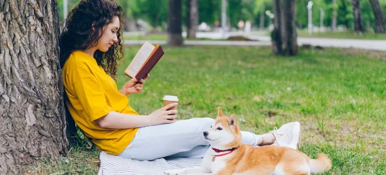 pexels-silverkblack-20897120.jpg Person reading a book and holding a coffee by a tree with a Shiba Inu dog beside them in a park
